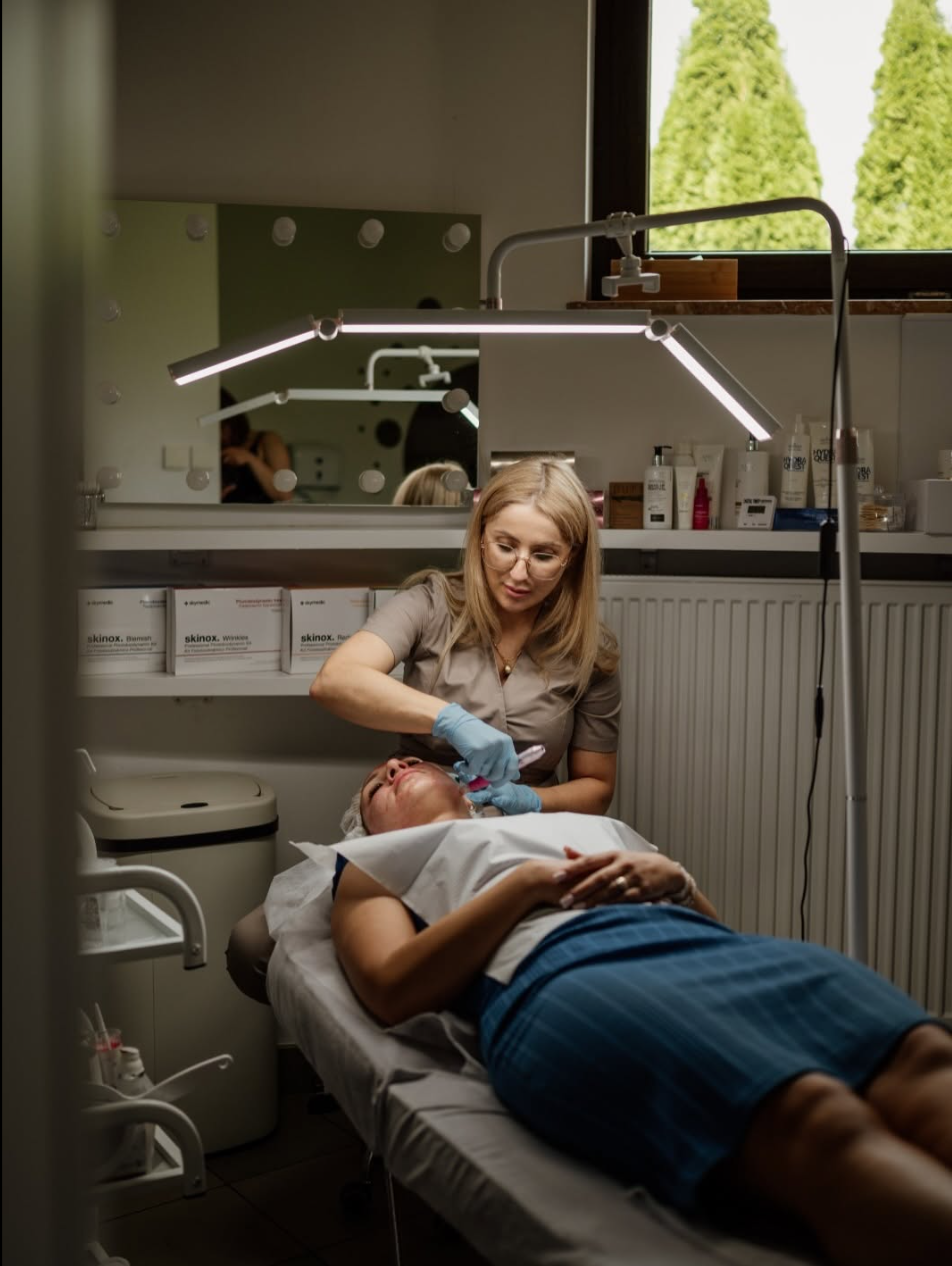 Pracuję przy zabiegu na twarzy klientki. Gabinet z lampą zabiegową i półką z kosmetykami.